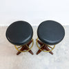 Two matching mid-century modern bar stools with round black leather seats, star-shaped dark wood bases, and brass circular ring accents, shown on a concrete floor against a white wall.
