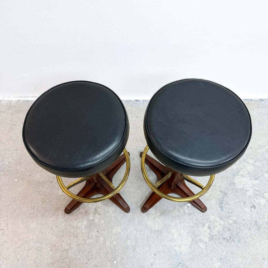 Two matching mid-century modern bar stools with round black leather seats, star-shaped dark wood bases, and brass circular ring accents, shown on a concrete floor against a white wall.