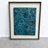 A framed abstract artwork featuring swirling, textured patterns in shades of teal, blue, and green, set within a white mat and a dark brown wooden frame, leaning against a white wall on a gray concrete floor.