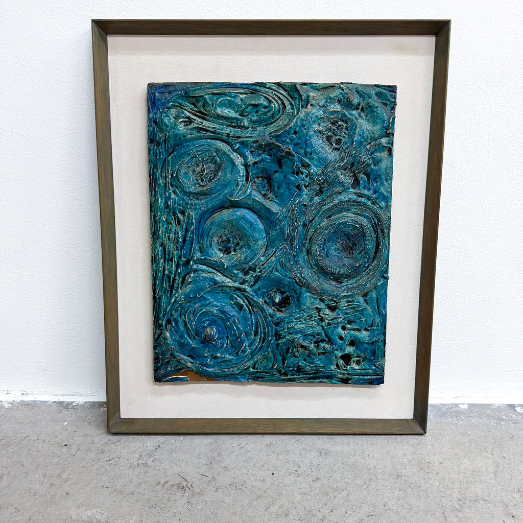 A framed abstract artwork featuring swirling, textured patterns in shades of teal, blue, and green, set within a white mat and a dark brown wooden frame, leaning against a white wall on a gray concrete floor.