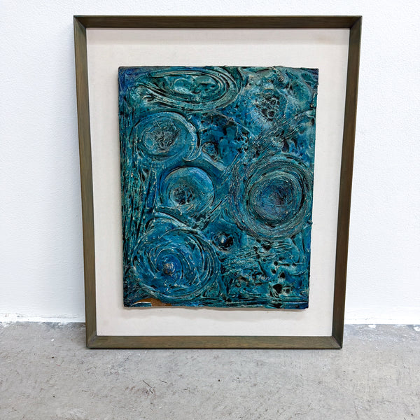 A framed abstract artwork featuring swirling, textured patterns in shades of teal, blue, and green, set within a white mat and a dark brown wooden frame, leaning against a white wall on a gray concrete floor.