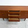 Mid-century modern wooden sideboard with three open drawers showing a light wood interior, photographed against a plain white wall and concrete floor.