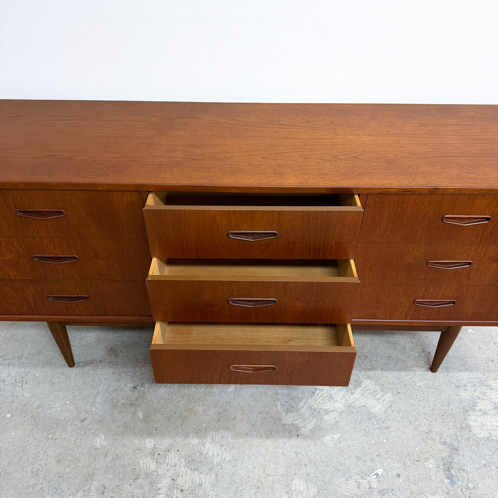 Mid-century modern wooden sideboard with three open drawers showing a light wood interior, photographed against a plain white wall and concrete floor.