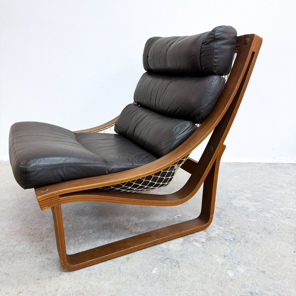 A mid-century modern lounge chair with black leather cushions and a curved wooden frame, photographed in a studio setting with a light gray concrete floor and white wall.