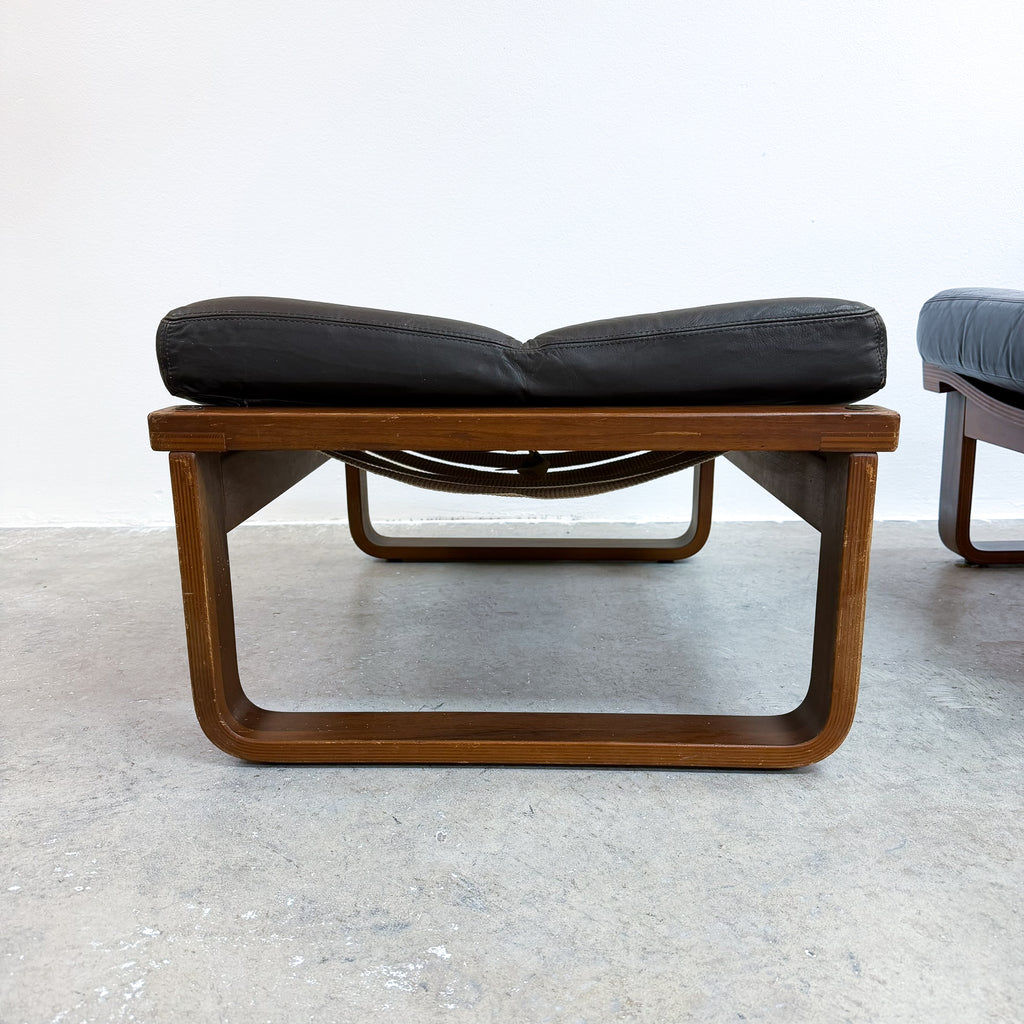 A mid-century modern lounge chair with a curved wooden frame and a black leather cushion, photographed in a minimalist room with a concrete floor and white wall.