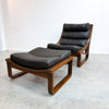 A mid-century modern wooden lounge chair with an attached matching footrest, upholstered in dark brown leather, set against a plain white wall and concrete floor.