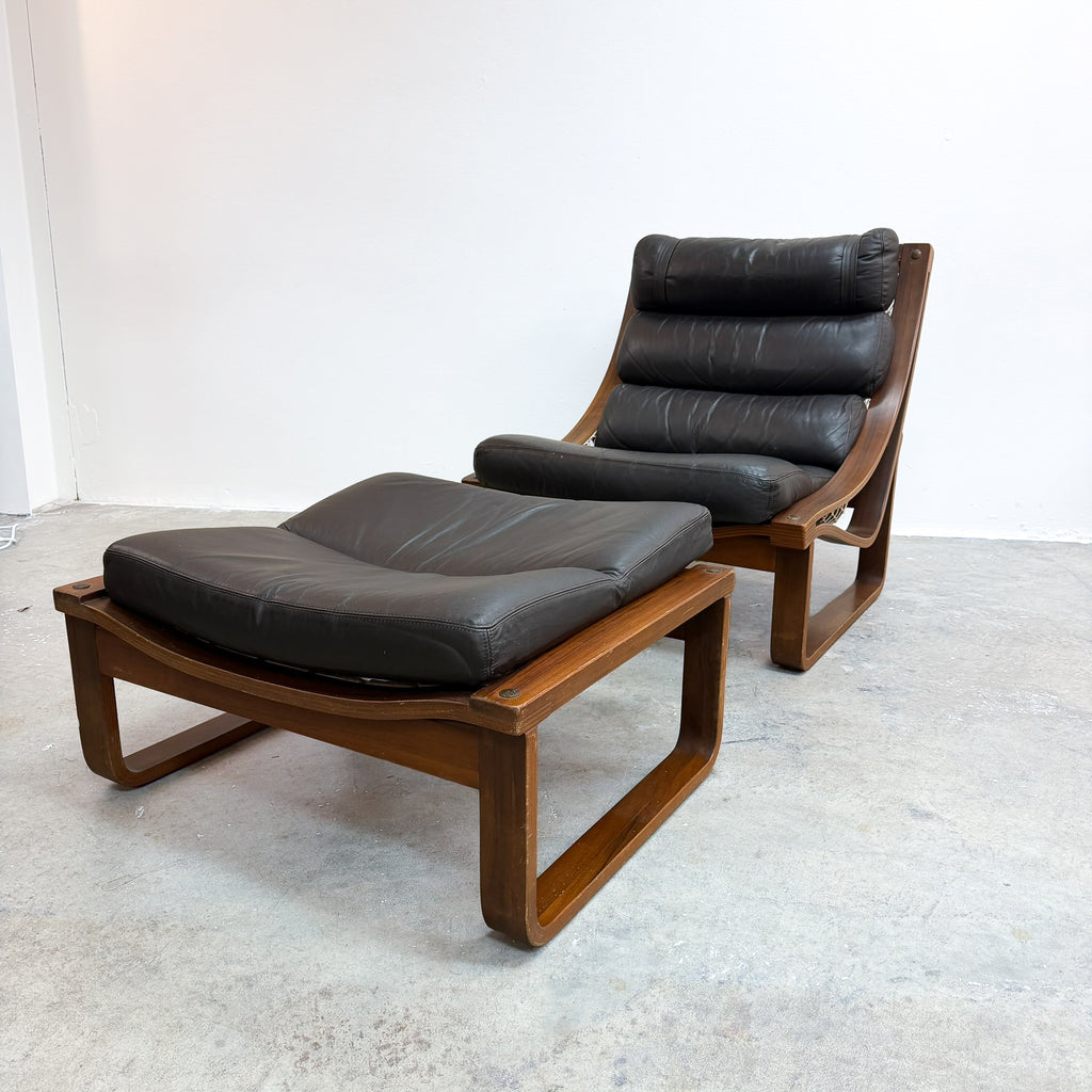 A mid-century modern wooden lounge chair with an attached matching footrest, upholstered in dark brown leather, set against a plain white wall and concrete floor.