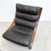 A mid-century modern lounge chair with dark brown leather cushions and a curved wooden frame, photographed on a concrete floor against a white wall.