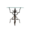 A unique table with a clear glass top and a dark brown, sculptural metal base resembling a stylized figure, set against a plain white wall and concrete floor.