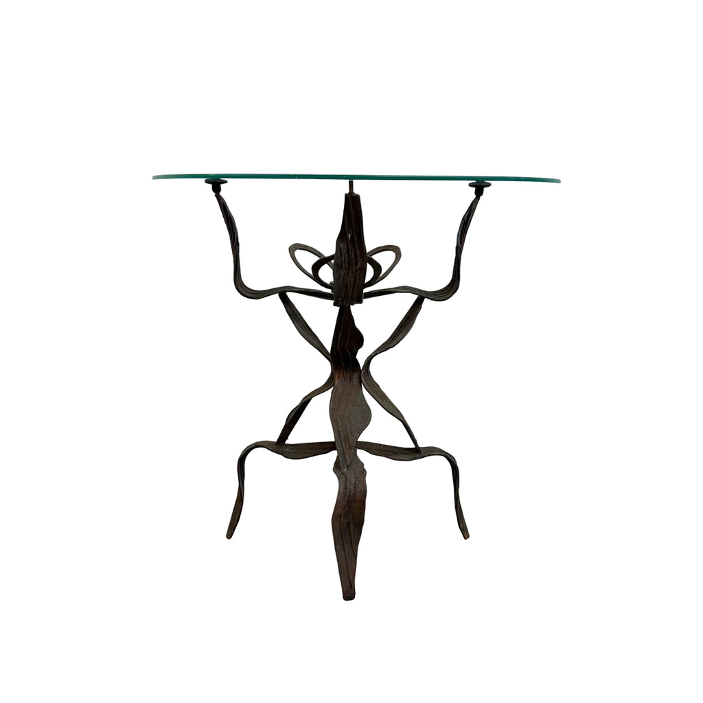 A unique table with a clear glass top and a dark brown, sculptural metal base resembling a stylized figure, set against a plain white wall and concrete floor.