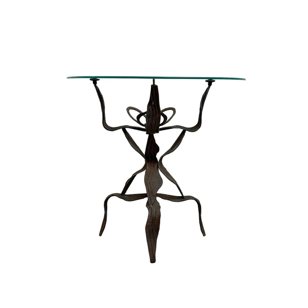 A unique table with a clear glass top and a dark brown, sculptural metal base resembling a stylized figure, set against a plain white wall and concrete floor.
