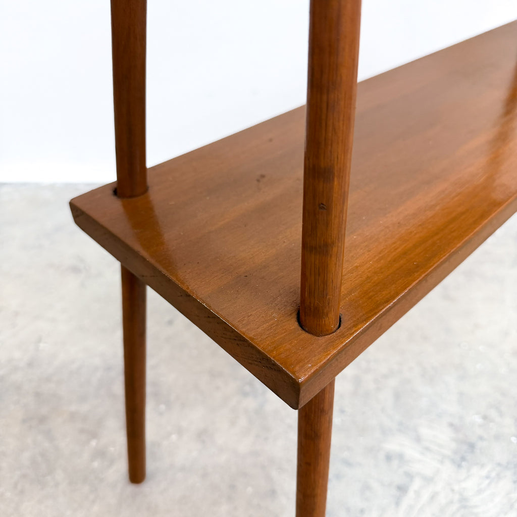 Close-up of a mid-century modern wooden bench with a warm brown finish, featuring a rectangular seat, vertical support posts, and tapered legs on a light gray concrete floor.