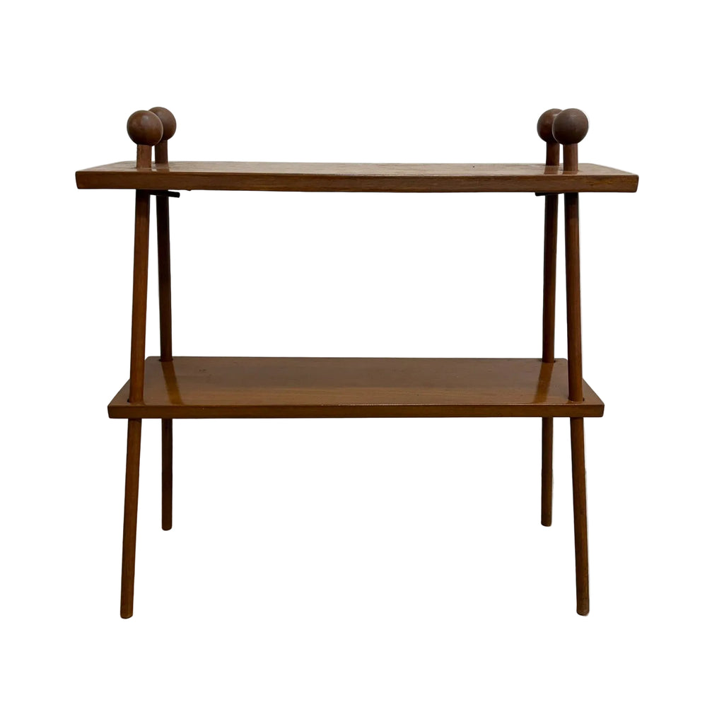 A mid-century modern wooden console table with two shelves, featuring tapered legs and decorative round finials on the top corners, set against a plain white wall and concrete floor.