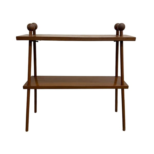 A mid-century modern wooden console table with two shelves, featuring tapered legs and decorative round finials on the top corners, set against a plain white wall and concrete floor.