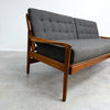 A mid-century modern wooden sofa with dark gray tufted upholstery, photographed in a minimalist studio setting with a concrete floor and white wall.
