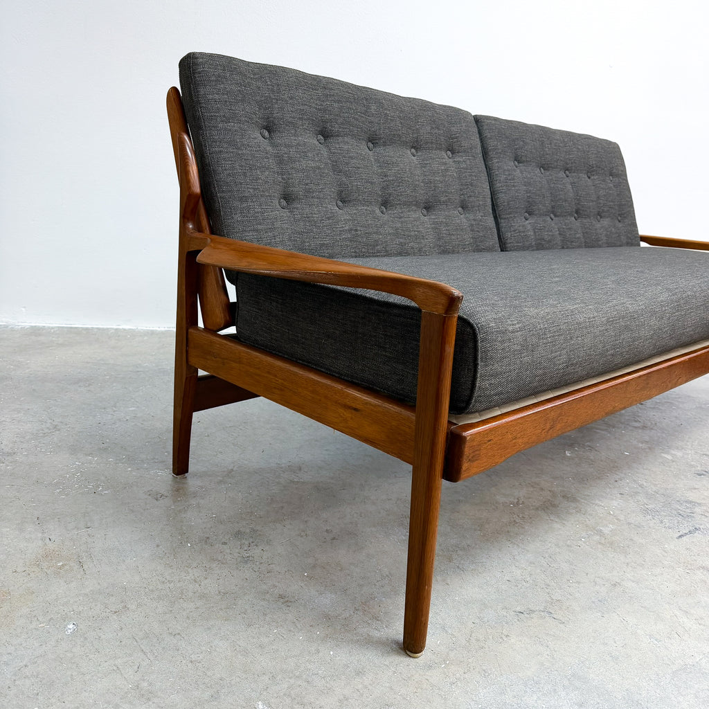A mid-century modern wooden sofa with dark gray tufted upholstery, photographed in a minimalist studio setting with a concrete floor and white wall.