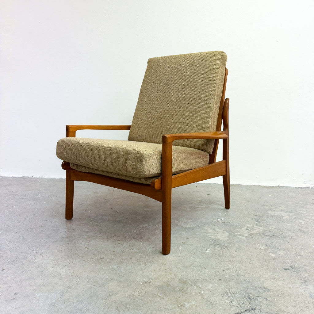 A mid-century modern armchair with a warm brown wooden frame and textured beige fabric cushions, set against a plain white wall and gray concrete floor.
