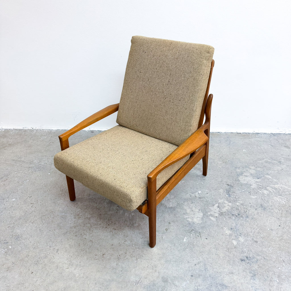A mid-century modern armchair with a warm medium-brown wooden frame and textured beige fabric upholstery, photographed against a plain white wall and gray concrete floor.