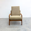 A mid-century modern armchair with a beige textured fabric cushion and a warm brown wooden frame, set against a plain white wall and gray concrete floor.