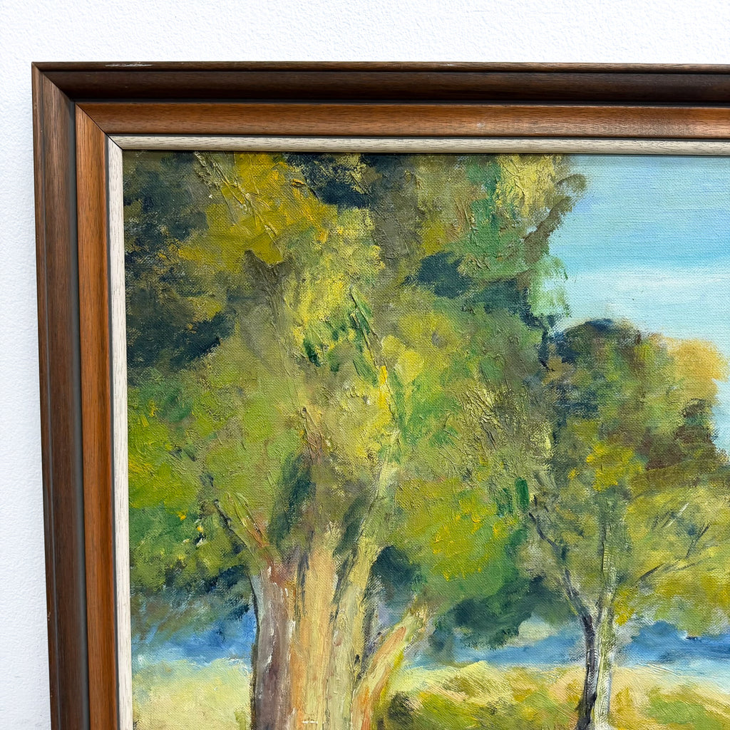 A framed landscape oil painting featuring green and yellow trees with blue sky, in a dark brown wooden frame against a white wall.