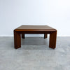 A mid-century modern wooden coffee table with a square top and thick block legs, shown on a concrete floor against a white wall.