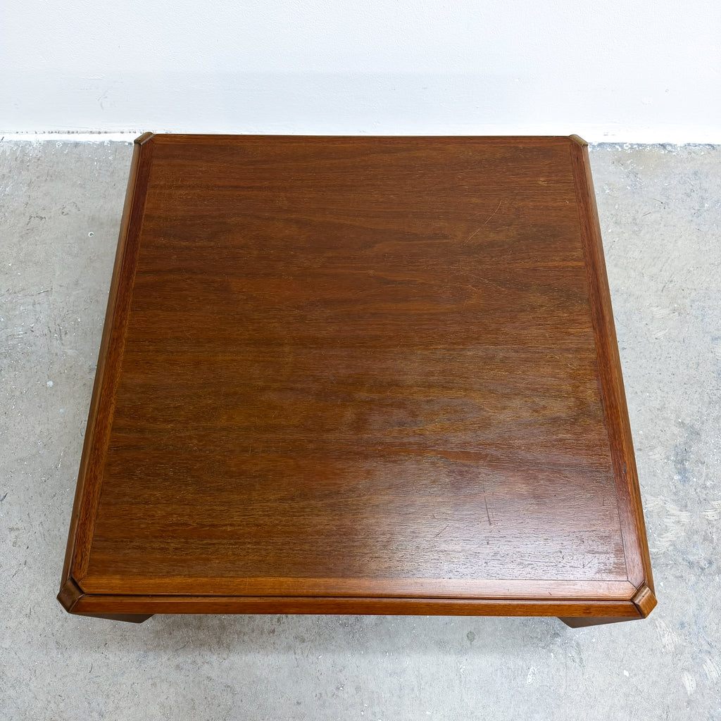 A square wooden side table or coffee table with a warm brown finish and beveled edges, photographed from directly above on a concrete floor against a white wall.