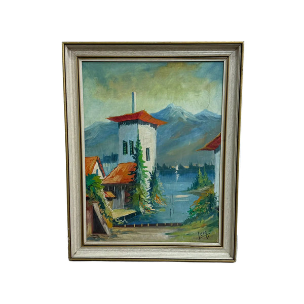 A framed landscape oil painting depicting a white tower with a red roof next to a lake, with mountains in the background, housed in a light-colored wooden frame and leaning against a white wall.