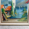 A framed landscape oil painting depicting a lakeside scene with a white building, red-orange roofs, green trees, and blue water, housed in a light-colored wooden frame.