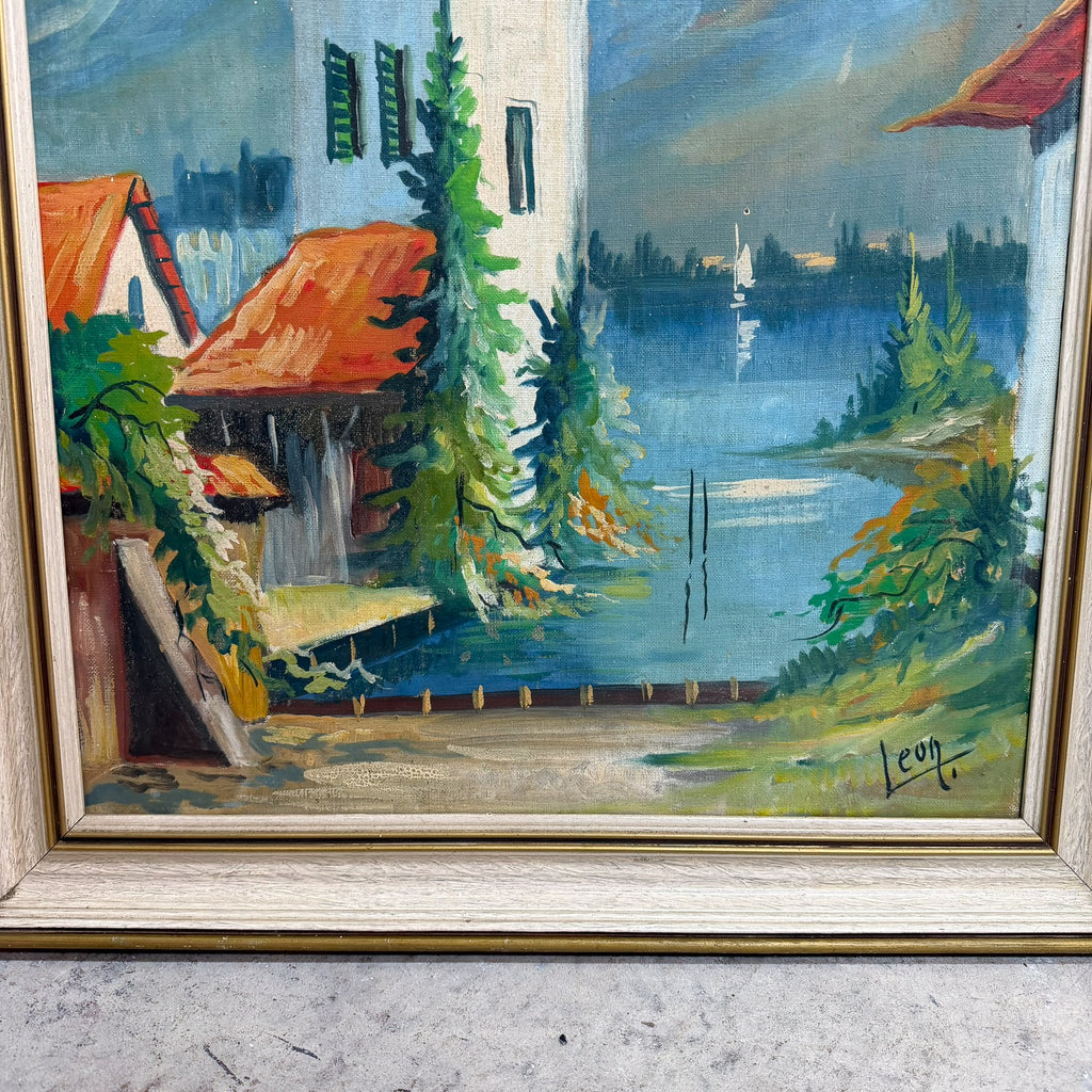 A framed landscape oil painting depicting a lakeside scene with a white building, red-orange roofs, green trees, and blue water, housed in a light-colored wooden frame.