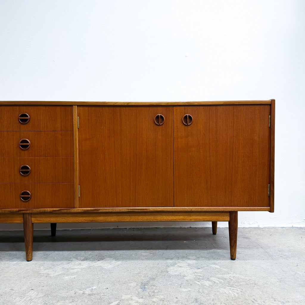 Mid-century modern wooden sideboard cabinet with a warm brown teak grain finish, featuring a drawer section on the left with circular pulls and two larger cabinet doors on the right with matching round pulls, set against a plain white wall and concrete floor.