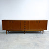 Mid-century modern wooden sideboard with a warm medium-brown finish, featuring multiple cabinet doors and drawers with dark round pulls, supported by tapered legs against a plain white wall and concrete floor.