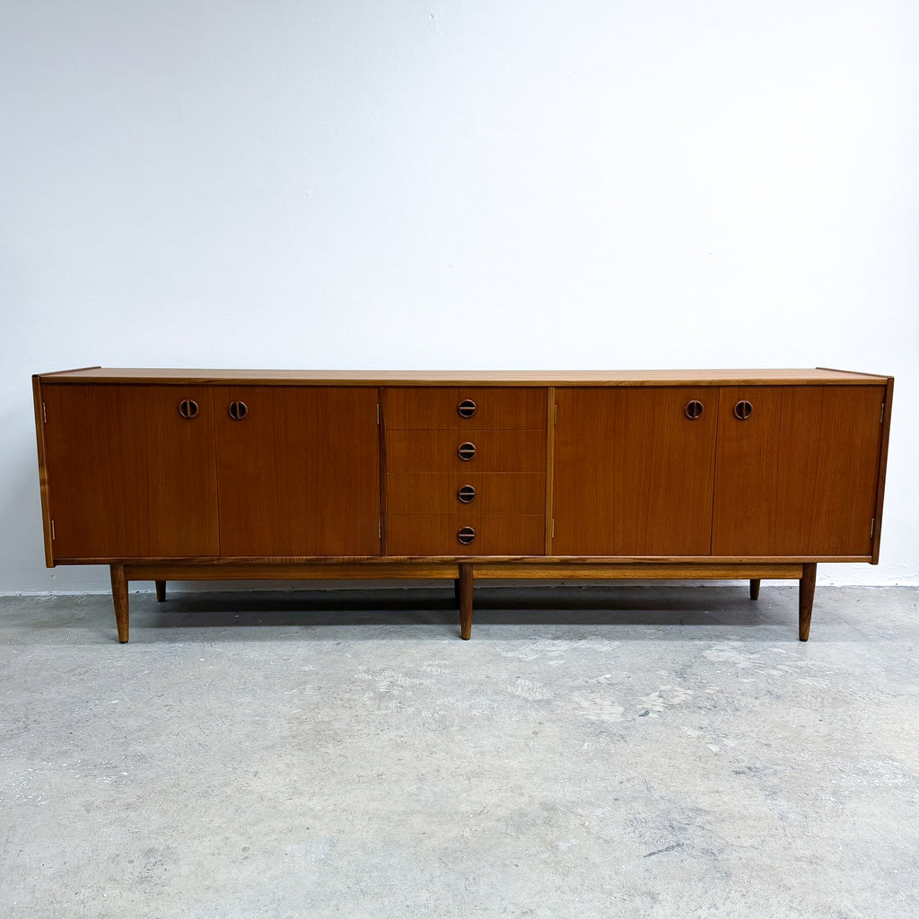 Mid-century modern wooden sideboard with a warm medium-brown finish, featuring multiple cabinet doors and drawers with dark round pulls, supported by tapered legs against a plain white wall and concrete floor.