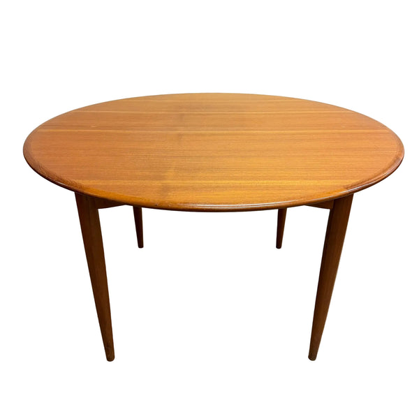 A mid-century modern round wooden dining table with tapered legs, shown on a concrete floor against a plain white wall.