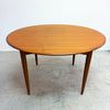 A mid-century modern round wooden dining table with tapered legs, shown on a concrete floor against a plain white wall.