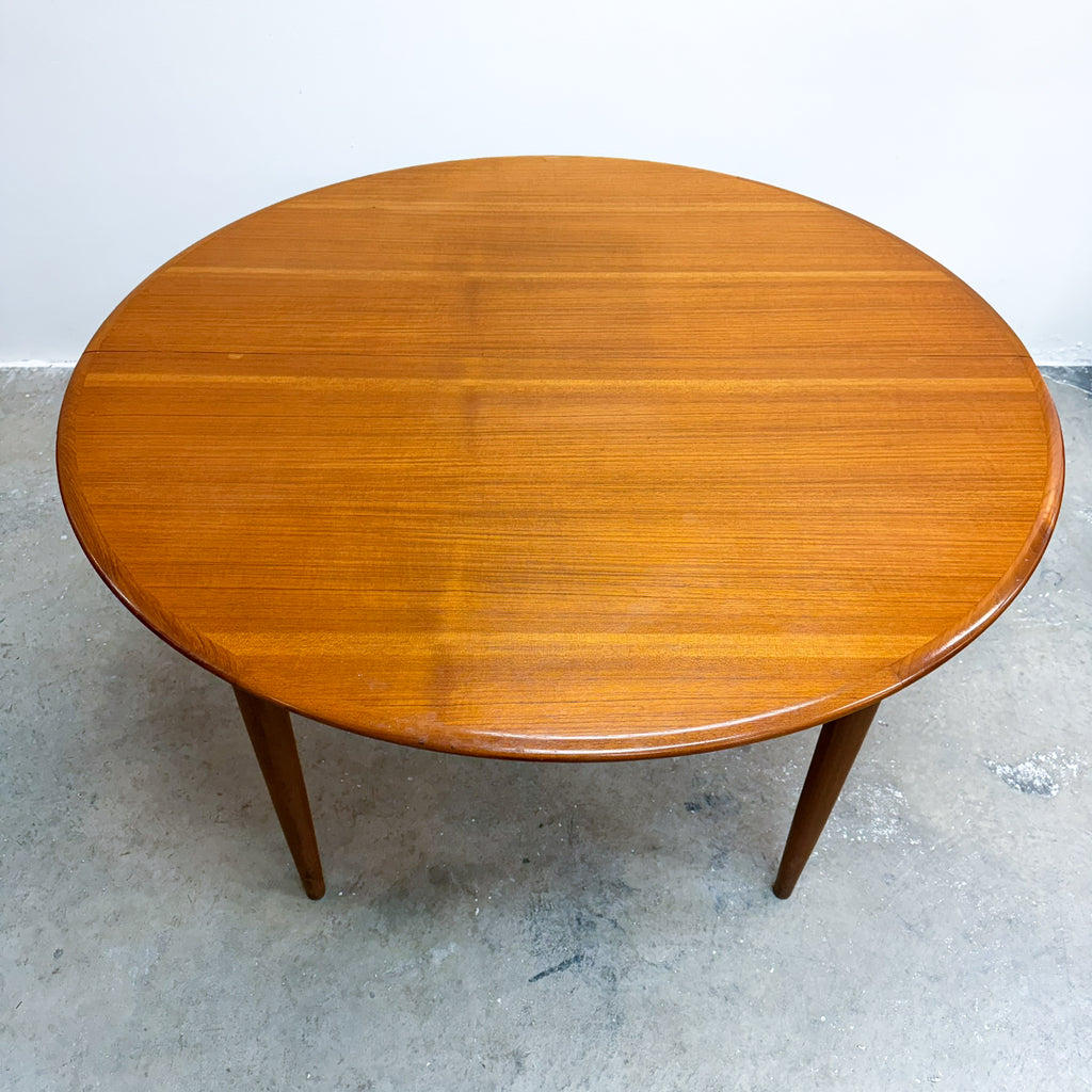 A round mid-century modern wooden dining table with a warm honey-amber finish and tapered legs, photographed on a concrete floor against a plain white wall.