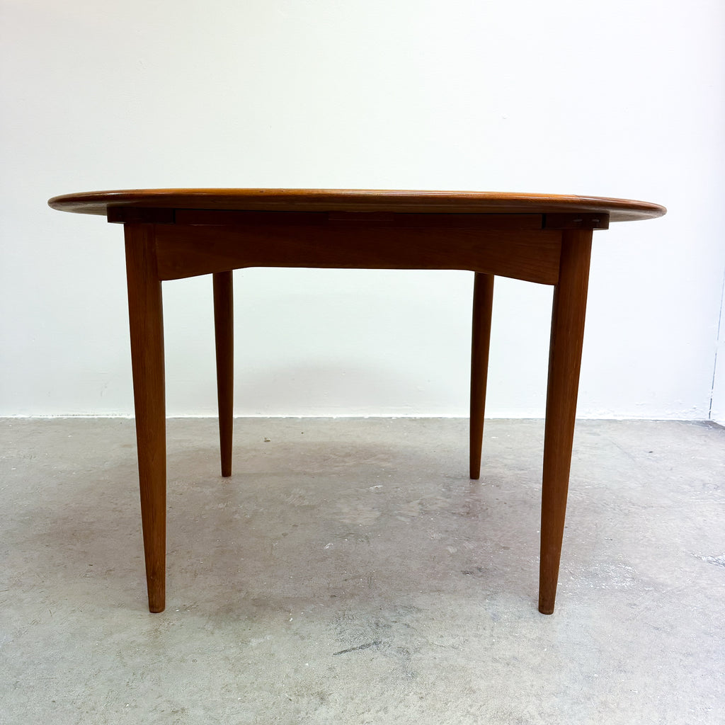 A mid-century modern teak dining table with a round top and tapered legs, shown against a plain white wall and concrete floor.