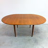 An oval mid-century modern wooden dining table with a warm medium-brown grain finish and tapered legs, set against a plain white wall and concrete floor.