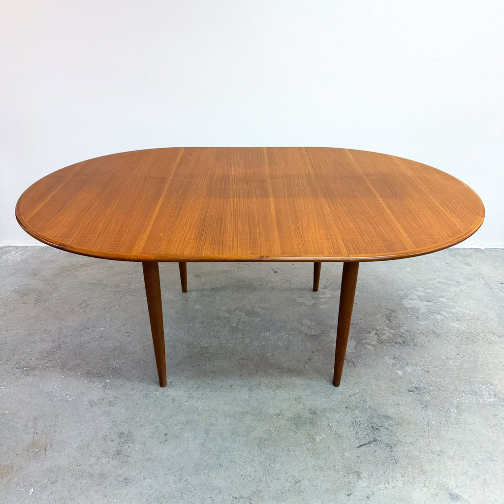 An oval mid-century modern wooden dining table with a warm medium-brown grain finish and tapered legs, set against a plain white wall and concrete floor.