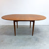 An oval mid-century modern wooden dining table with tapered legs, shown on a concrete floor against a plain white wall.