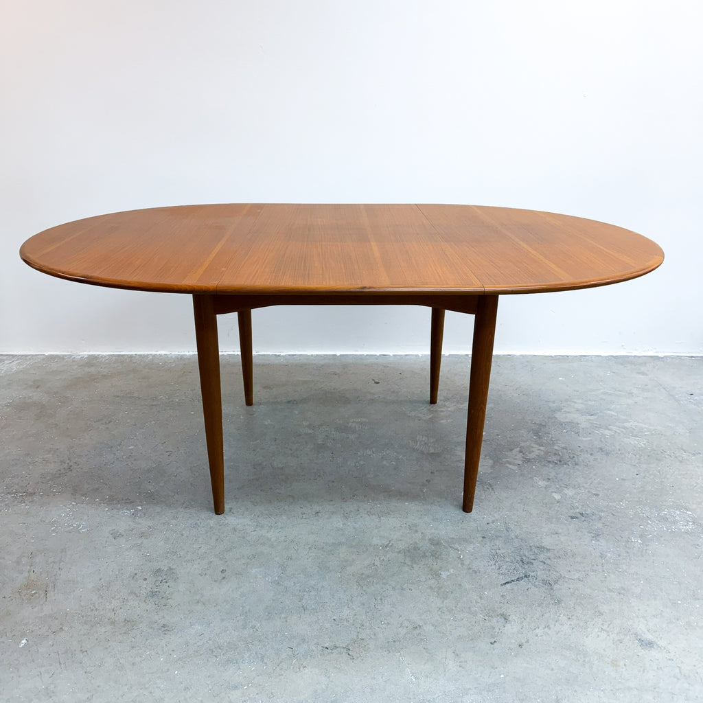 An oval mid-century modern wooden dining table with tapered legs, shown on a concrete floor against a plain white wall.