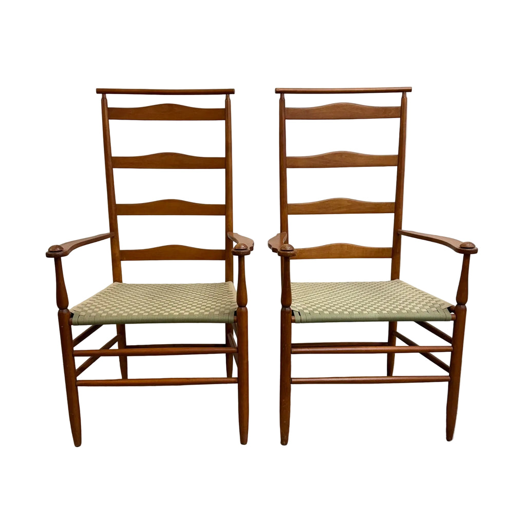 Two matching mid-century modern wooden dining chairs with ladder-back designs, armrests, and woven seat cushions, set against a plain white wall and concrete floor.