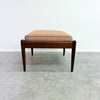 A mid-century modern wooden bench or ottoman stool with a square cushion upholstered in a light tan fabric, set against a plain white wall and gray concrete floor.