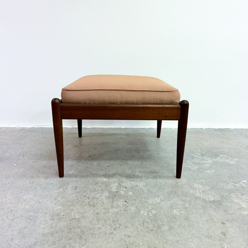 A mid-century modern wooden bench or ottoman stool with a square cushion upholstered in a light tan fabric, set against a plain white wall and gray concrete floor.