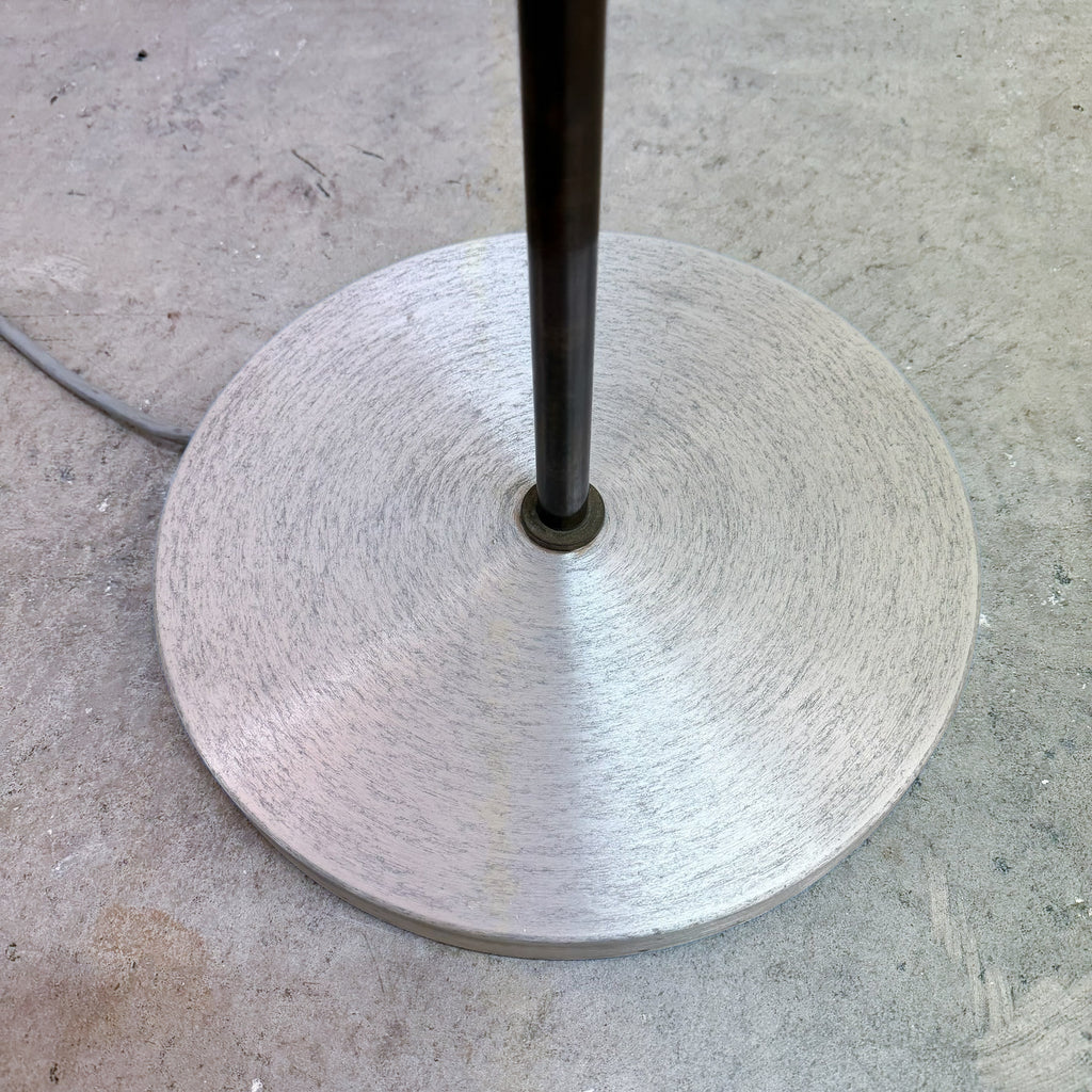 A brushed metal circular base with a central dark metal pole, photographed on a concrete floor.