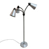 A mid-century modern three-light floor lamp with a black metal frame and silver conical metal shades, standing on a concrete floor against a white wall.