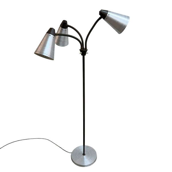 A mid-century modern three-light floor lamp with a black metal frame and silver conical metal shades, standing on a concrete floor against a white wall.