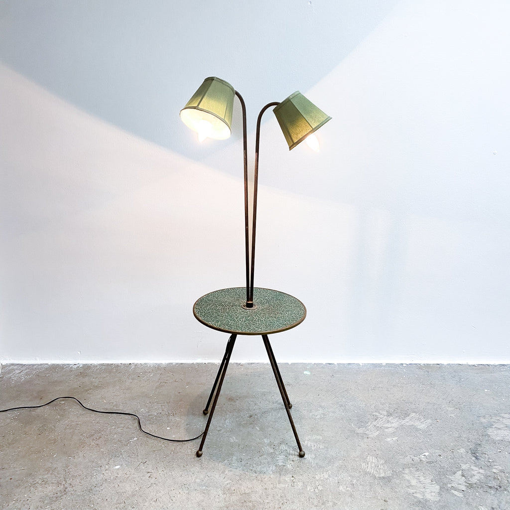 Floor lamp with two green shades on a round metal table against a white wall.