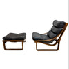 A mid-century modern lounge chair with an ottoman, featuring black leather cushions and a curved wooden frame, set against a plain white wall and concrete floor.