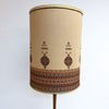 A cylindrical fabric lampshade with a beige linen-like base and traditional geometric embroidery in brown, red, and black, shown on a lamp stand against a plain white background.