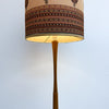 A vintage-style floor lamp with a tapered beige fabric shade featuring a detailed brown, black, and red geometric pattern, supported by a slender brown wooden stem against a plain white wall.